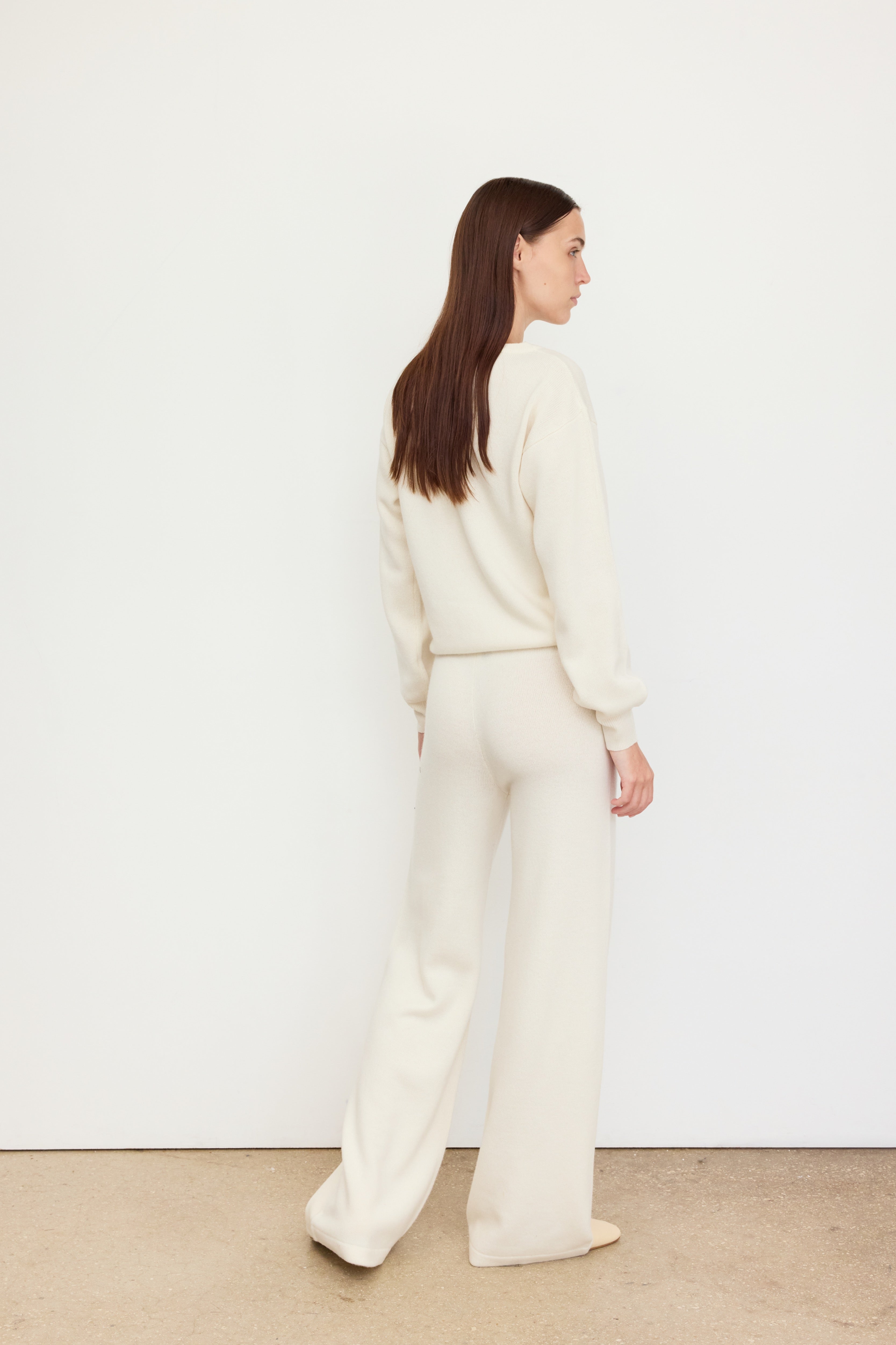 A woman stands in a relaxed pose with her back facing the camera, her head turned slightly to the side.
She is wearing an off-white knit set made of 100% Carrigi cashmere, featuring a long-sleeve sweater and wide-leg pants.
Her long, straight brown hair falls naturally down her back. The minimal setting with a white wall and neutral floor highlights the soft drape and luxurious texture of the cashmere outfit.