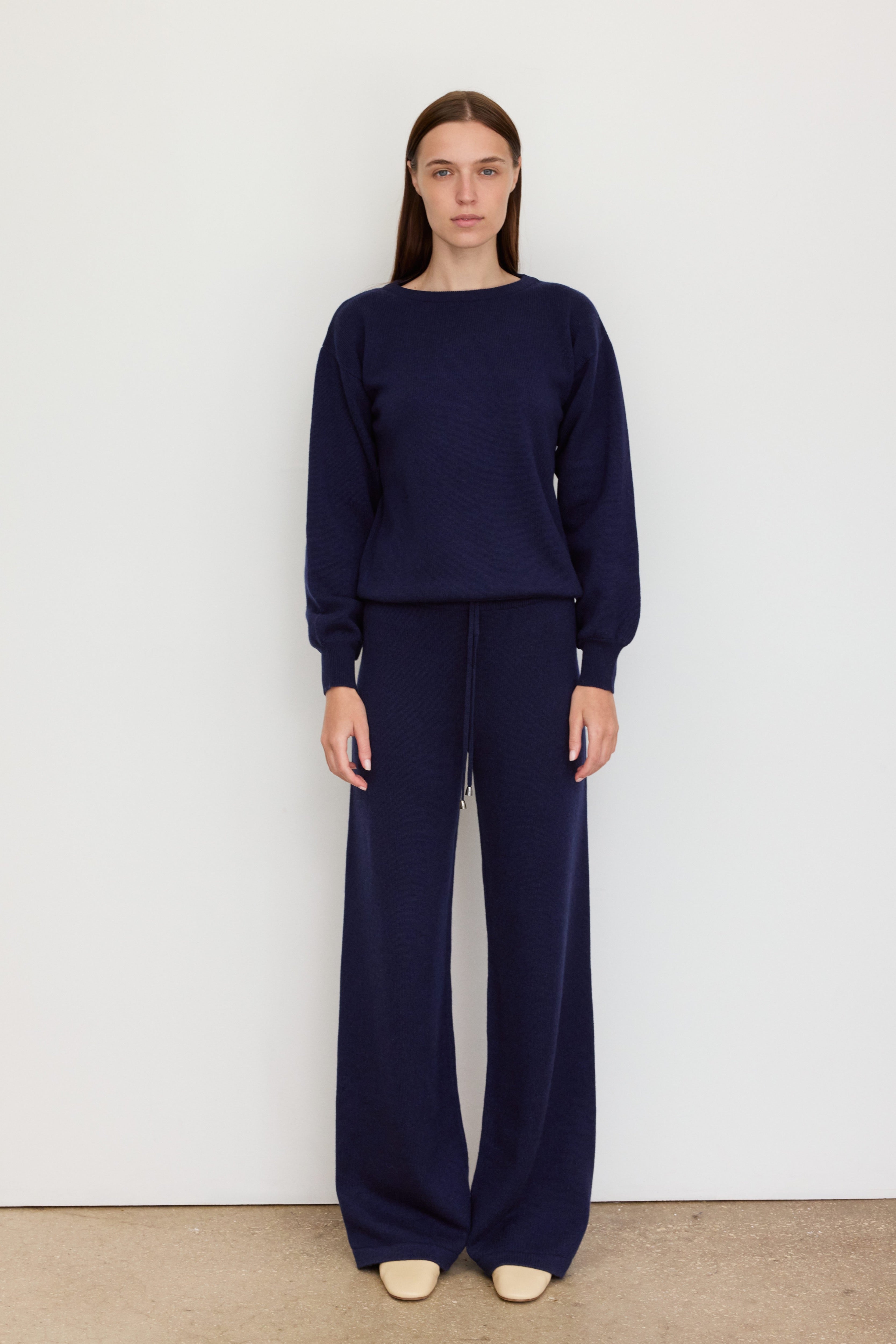 A woman stands facing the camera against a plain white wall. She wears a dark navy knit ensemble consisting of a crewneck sweater and wide-leg drawstring pants. Her hair is straight and tucked behind her ears, and her expression is calm and composed. She pairs the outfit with cream-colored slip-on shoes. The minimalist background and neutral flooring emphasize the clean lines and relaxed elegance of the outfit.