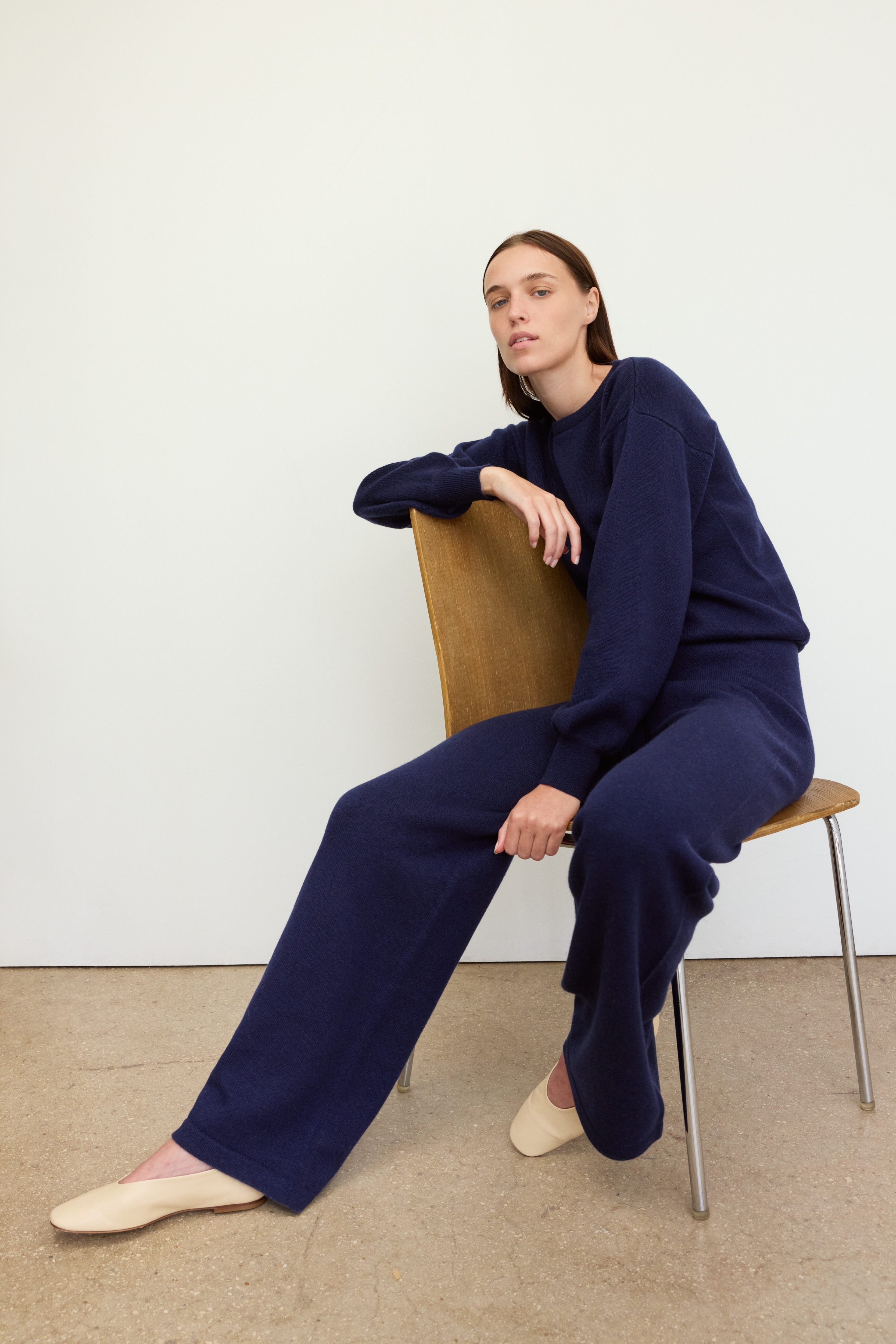 A woman with straight brown hair sits on a simple wooden chair against a white wall. She is dressed in a loose-fitting, dark navy knit outfit 'Verona Sweater Navy,with wide-leg pants and a matching sweater. Her posture is relaxed and confident, with one arm resting on the back of the chair and one leg extended forward. She wears cream-colored slip-on shoes, and the setting is minimal with a neutral-toned floor and no distractions in the background.