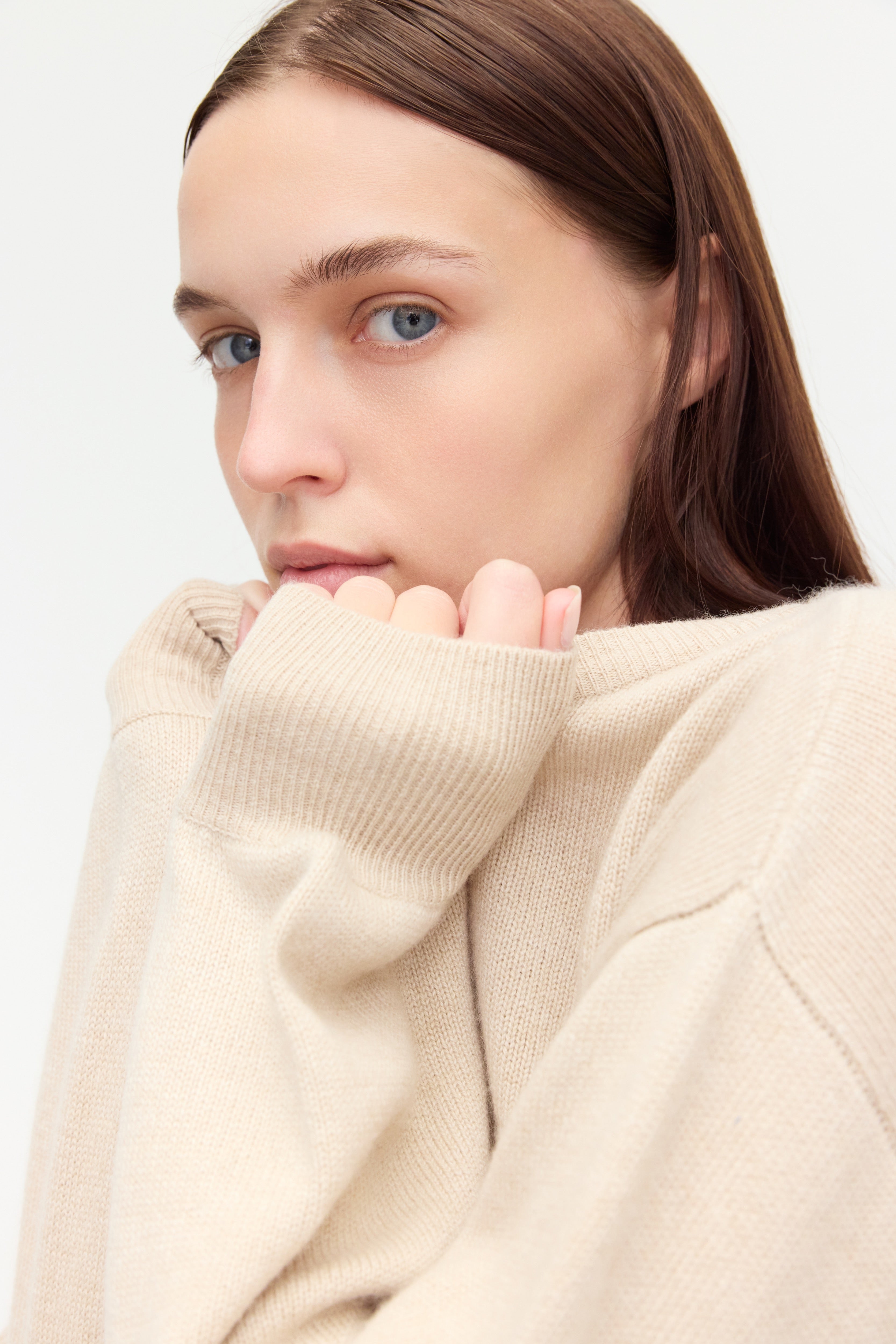 A close-up portrait of a woman with blue eyes and long brown hair, wearing a soft beige knit sweater made of 100% cariggi cashmere.
She gazes softly to the side with a contemplative expression, gently holding the collar of her sweater near her face. The image emphasizes the cozy texture of the fabric and the natural, serene mood of the composition.