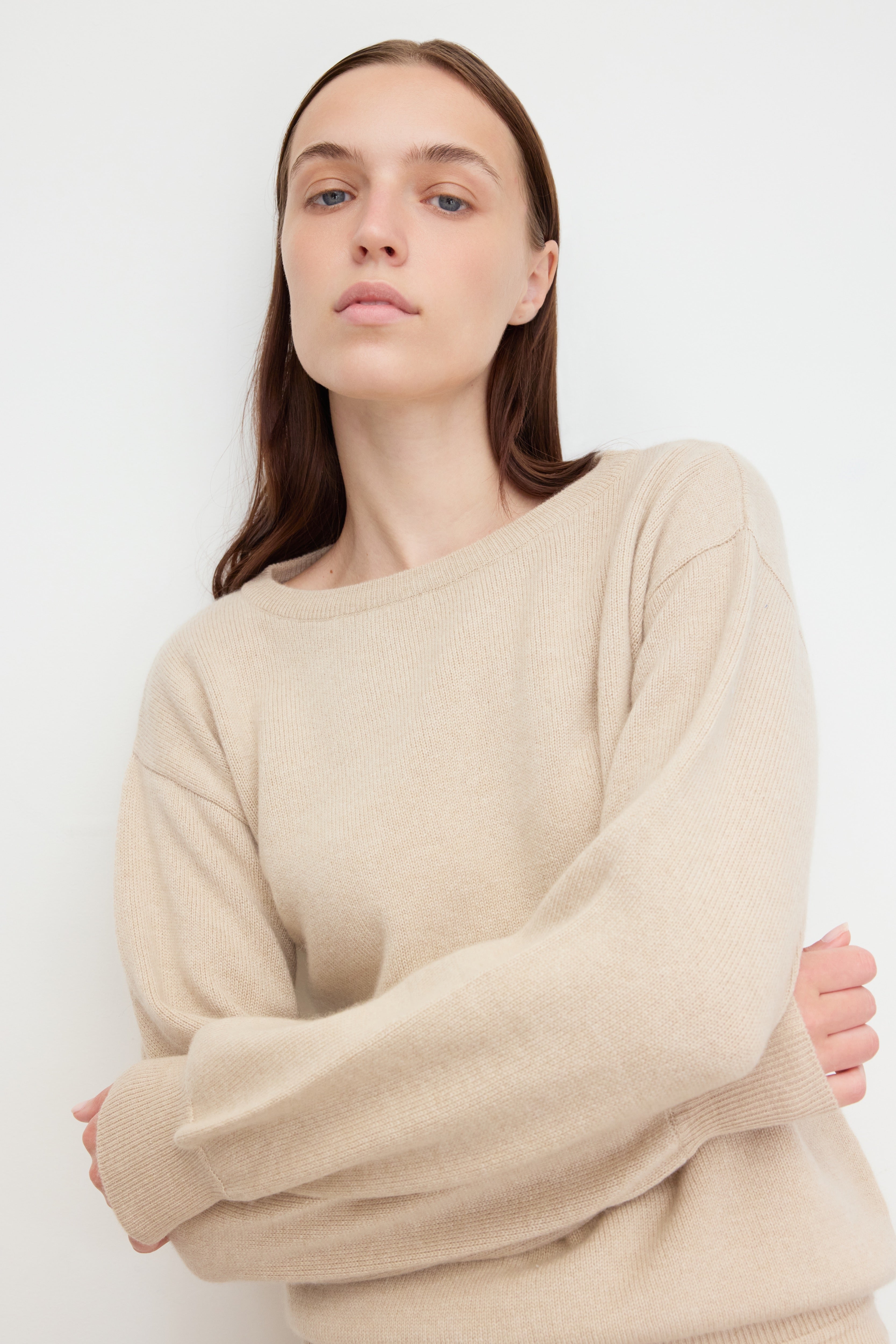 A woman with long, straight brown hair poses confidently against a plain white background.
She is wearing a beige knit sweater made of 100% cariggi cashmere.
With relaxed sleeves, her arms loosely crossed in front of her.
Her expression is calm and self-assured, and the close-up framing emphasizes the minimal, natural aesthetic of the outfit and setting.