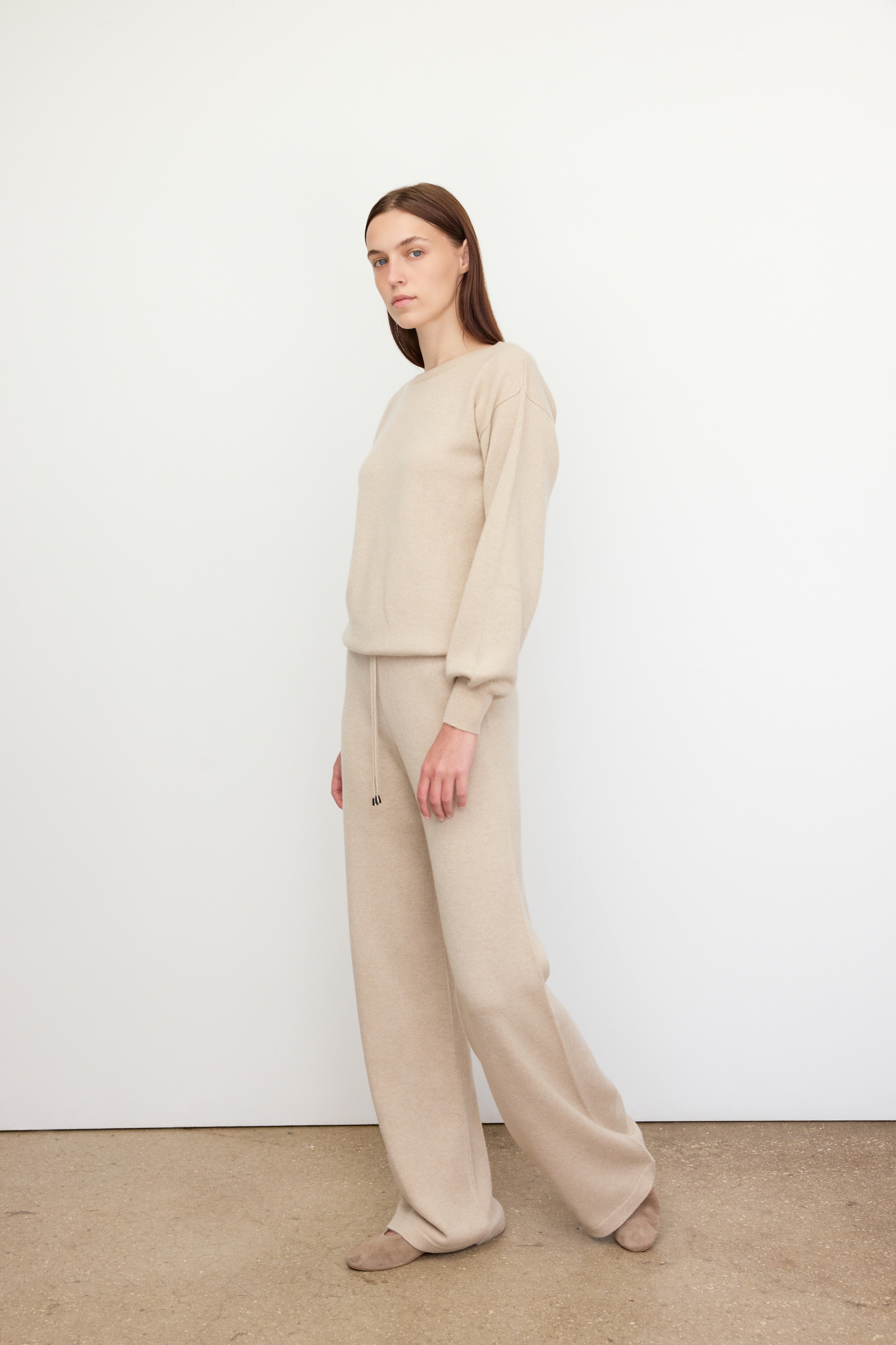 A woman stands in a relaxed pose against a plain white wall, wearing a coordinated beige knit outfit. The ensemble includes a long-sleeve sweater and wide-leg drawstring pants made from a soft 100% cariaggi cashmere. Her hands are by her sides, and she gazes slightly to the left with a calm expression. She completes the look with matching beige slippers, and the minimalist background emphasizes the cozy, monochromatic aesthetic.