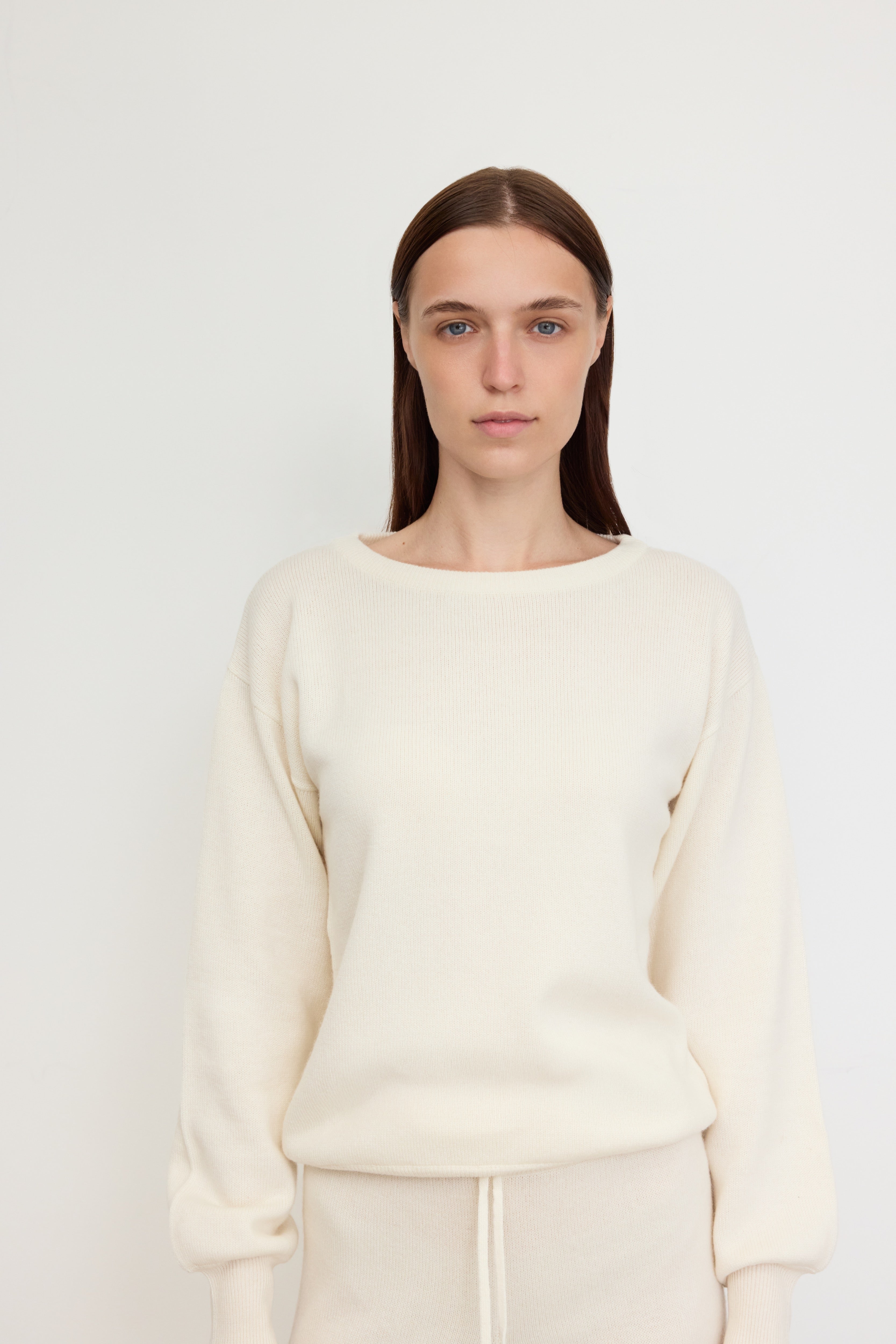A woman stands facing the camera against a plain white background, wearing a coordinated off-white knit set made of 100% Carrigi cashmere. 
The outfit includes a long-sleeve sweater with a relaxed fit and drawstring pants. 
Her hair is straight and tucked behind her ears, and her expression is calm and composed. 
The clean backdrop highlights the luxurious texture and simplicity of the cashmere ensemble.