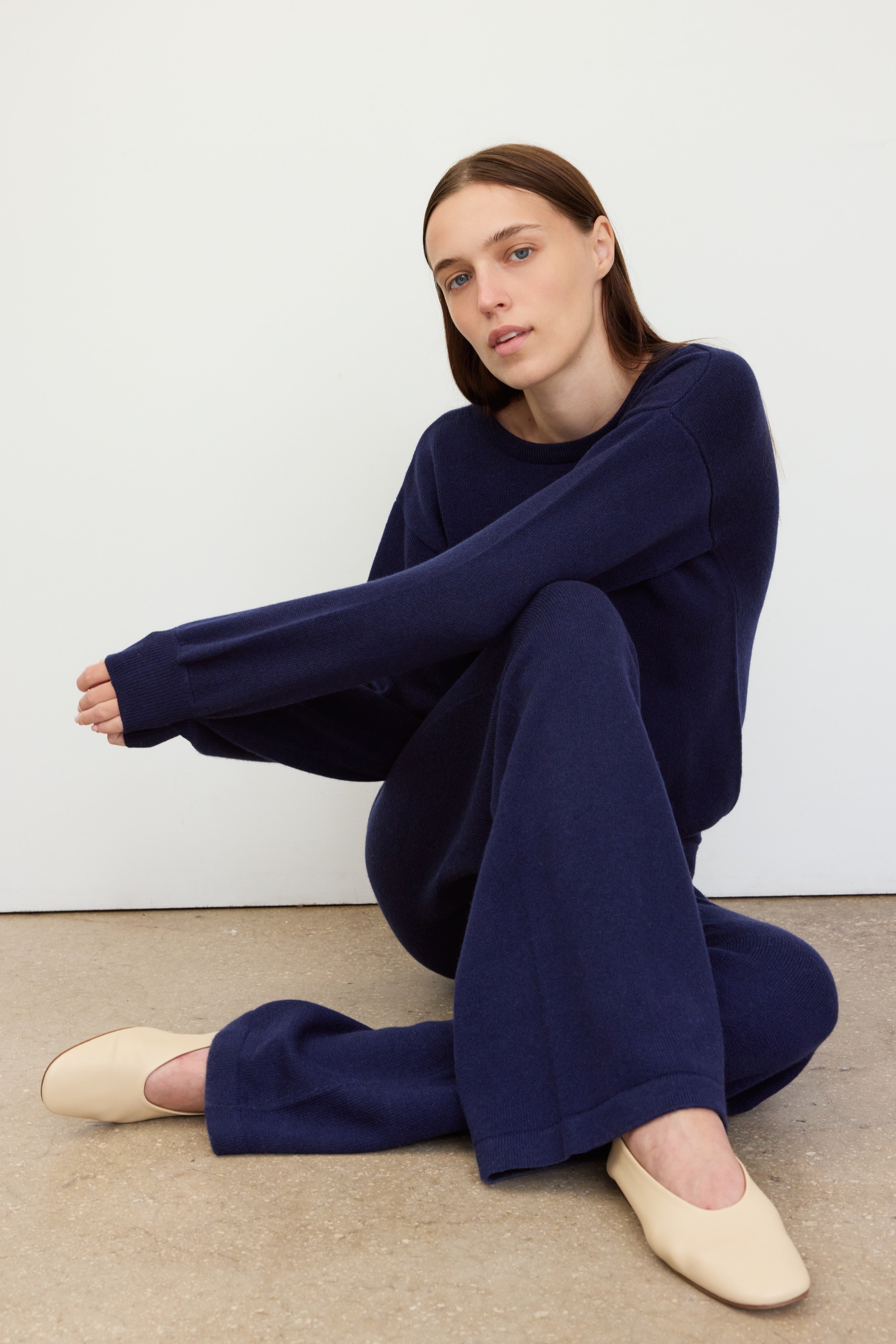 A woman with straight brown hair sits on the floor against a light-colored wall. 
She is wearing a relaxed, navy blue knit set with flared pants and long sleeves, paired with cream-colored slip-on shoes.
Her pose is casual and introspective, with one arm resting on her knee and the other extended across her legs. 
The background is minimal, emphasizing the simplicity and softness of the outfit.