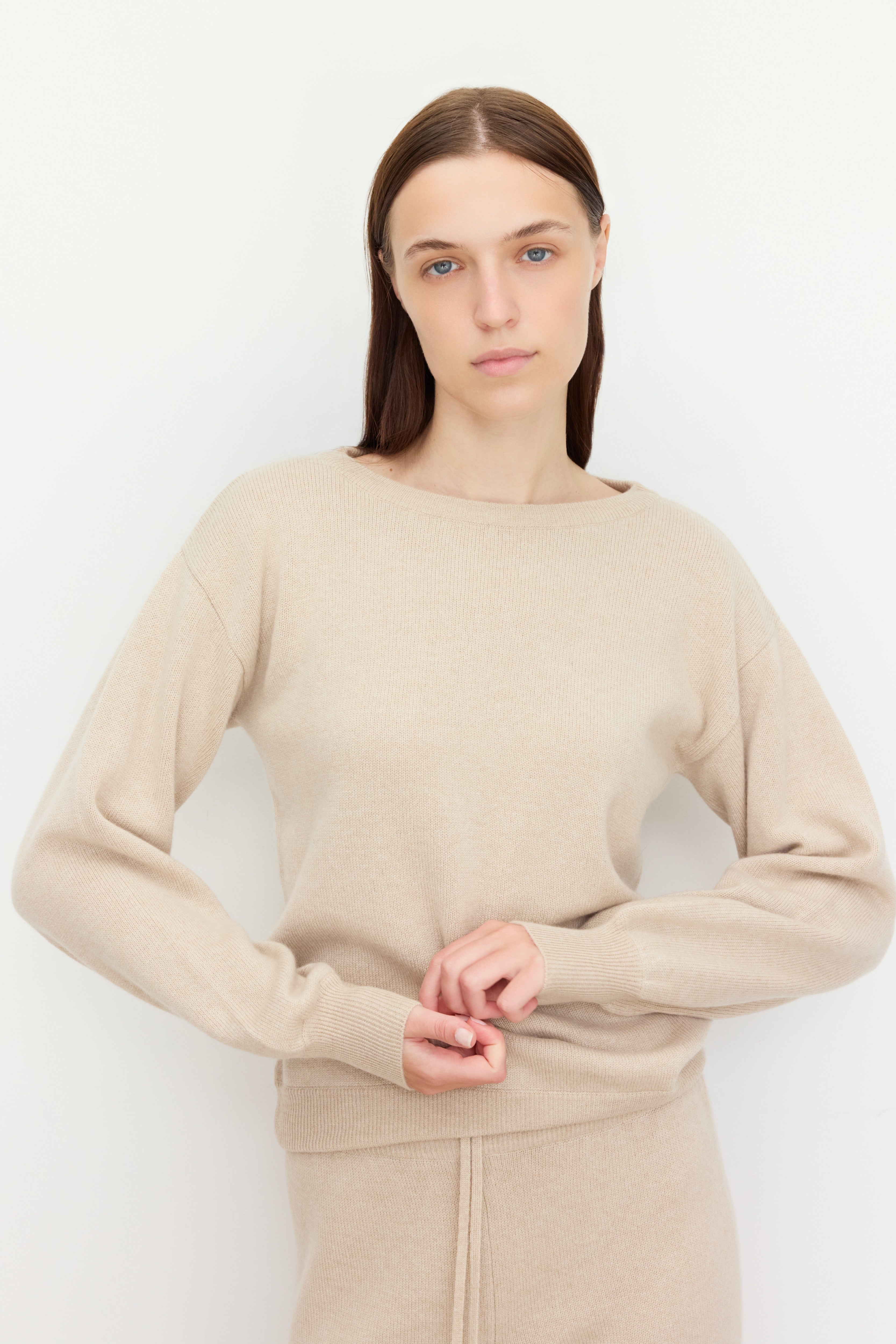 A woman with straight brown hair stands against a light-colored wall, looking directly at the camera with a calm expression. She is wearing a soft beige knit set consisting of a long-sleeve sweater and matching drawstring pants made of  100% cariaggi cashmere. 
Her hands are gently clasped at her waist, subtly adjusting the hem of her top. The neutral tones and minimal backdrop highlight the cozy texture and simplicity of the outfit.